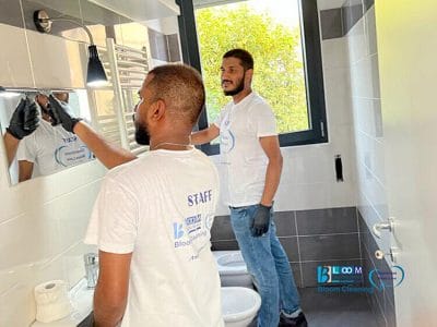 Due operai della BloomCleaning, che indossano camicie bianche e guanti, stanno pulendo un bagno con piastrelle grigie e bianche, un water, un bidet e una finestra. Uno sta lucidando lo specchio mentre l'altro osserva. Pulizia appartamenti Inganni Milano assicura che ogni dettaglio brilli.