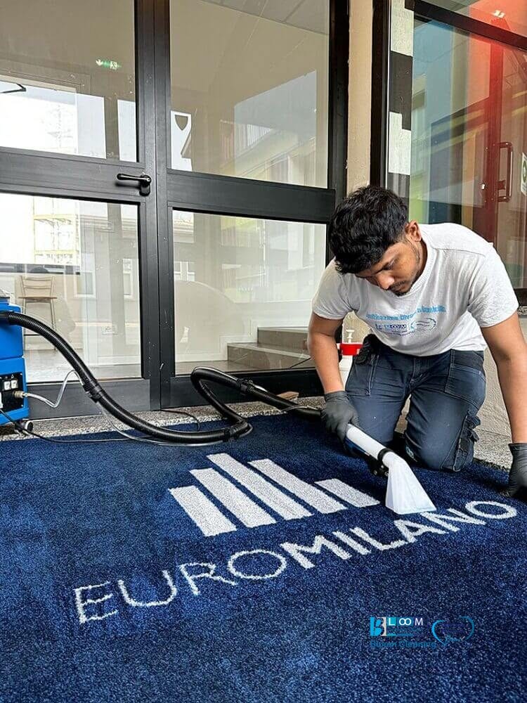 Un uomo pulisce un tappeto blu davanti a un edificio di Milano utilizzando il metodo Iniezione/Estrazione.