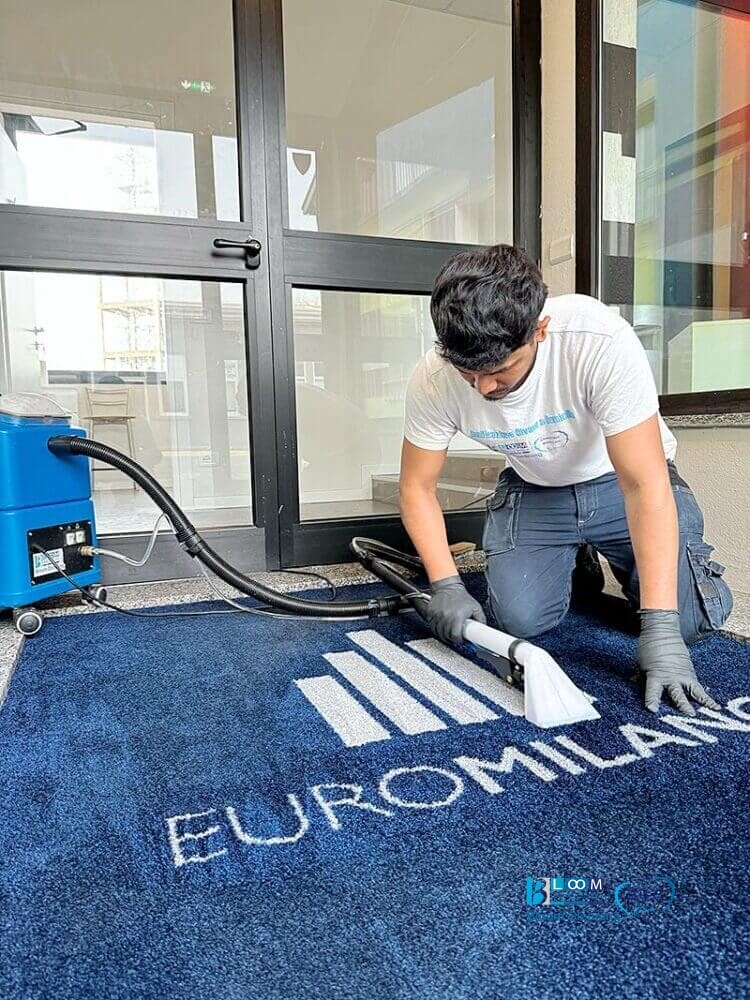 Un uomo che pulisce un tappeto blu davanti a un edificio utilizzando il metodo dell'iniezione/estrazione a Milano.