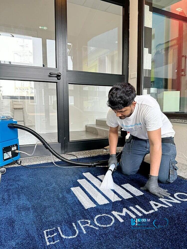 Un uomo che pulisce un tappeto blu davanti a un edificio utilizzando il metodo dell'iniezione/estrazione a Milano.