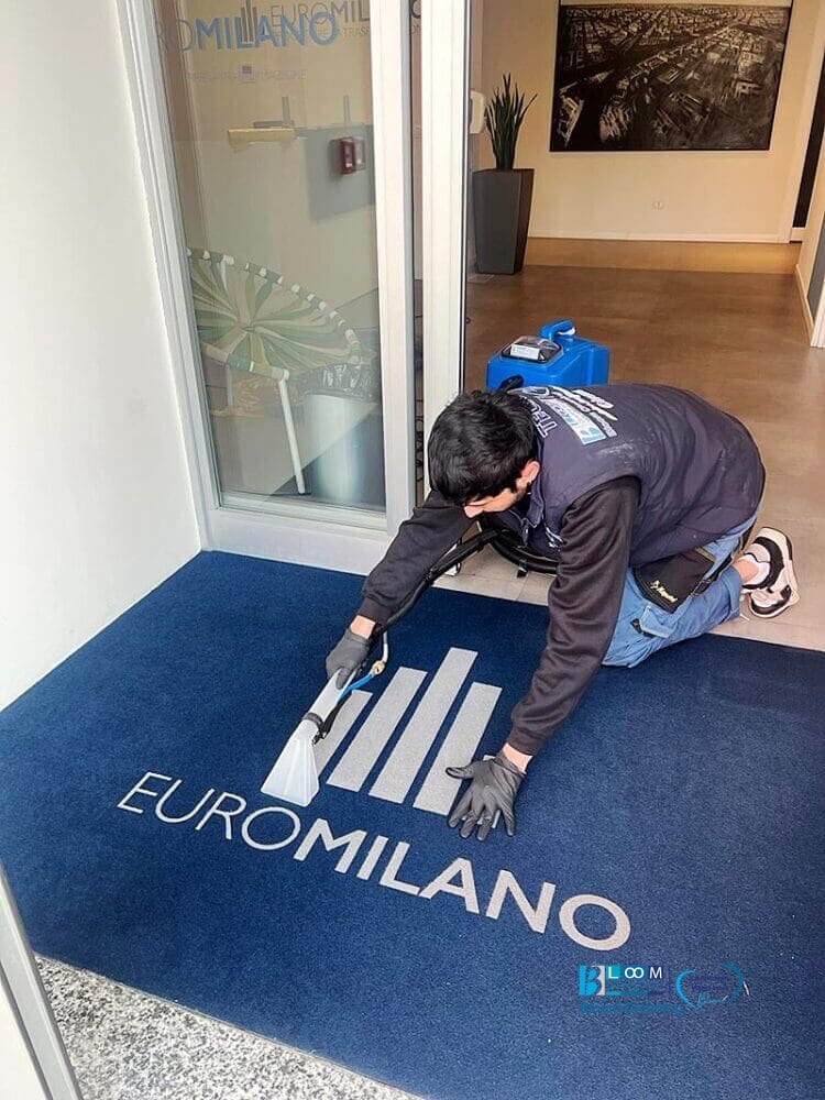 Un uomo che pulisce un tappeto blu davanti a un edificio utilizzando il metodo dell'iniezione/estrazione a Milano.