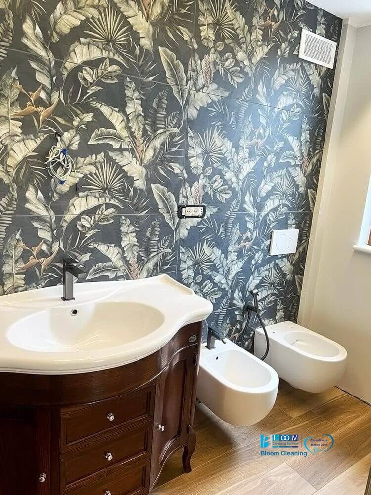 Un bagno moderno con carta da parati dal design botanico, caratterizzato da un classico lavabo in legno e sanitari bianchi, pulito da Bloom Cleaning.