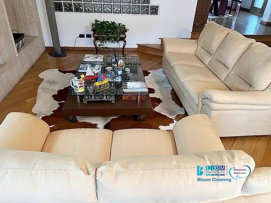 Un accogliente soggiorno dal design contemporaneo caratterizzato da un divano in Alcantara color crema, un divanetto coordinato e un tavolino in legno ornato con vari oggetti decorativi, posto su un tappeto in pelle di mucca.