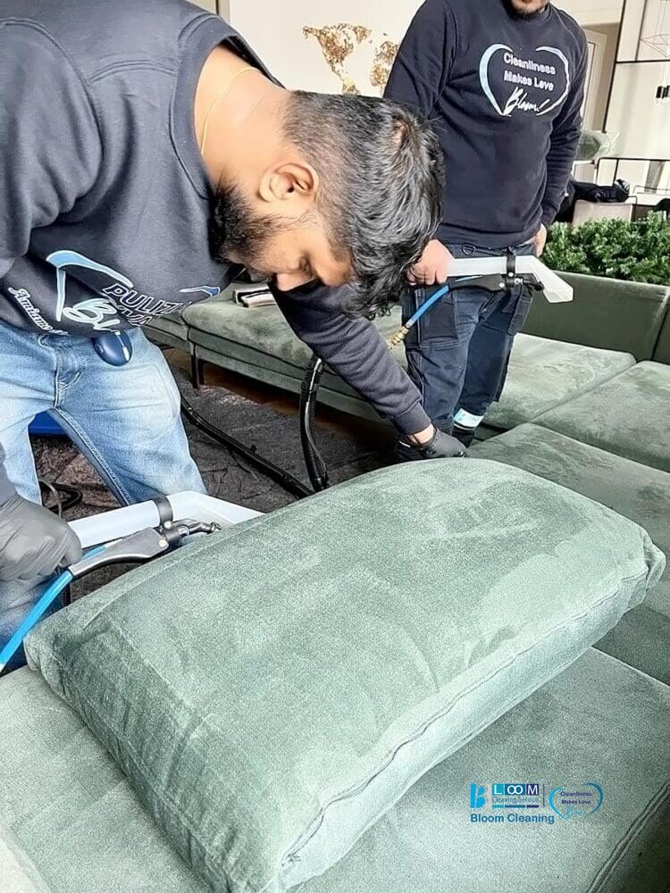 Due persone puliscono un cuscino imbottito verde con un aspirapolvere automatico in una stanza ben illuminata.