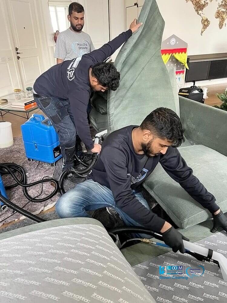 Tre persone puliscono un divano con attrezzature professionali in un ambiente domestico con tiraggio automatico.