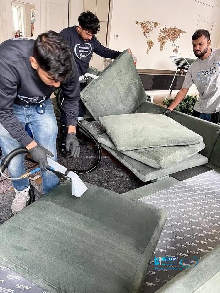 Tre uomini puliscono professionalmente un divano verde, uno dei quali utilizza uno strumento di pulizia Bozza automatica sui cuscini del divano.