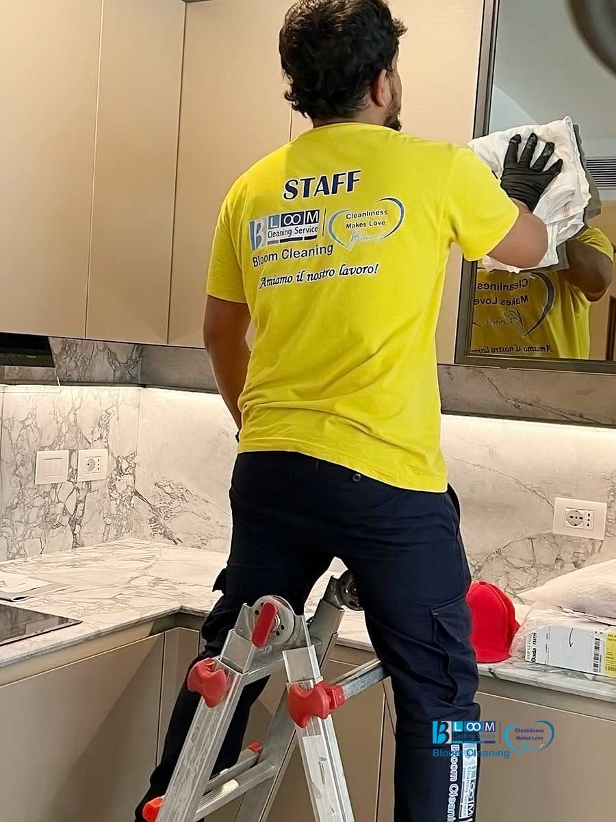 Professionista del servizio di pulizia impegnato nella pulizia appartamenti milano, pulisce il mobile della cucina stando in piedi su una scala.