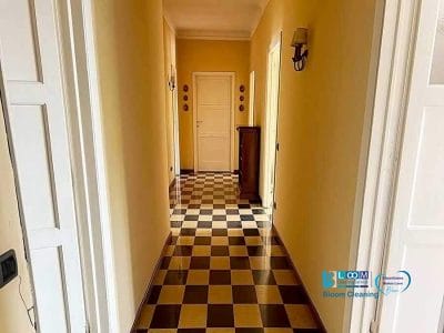 Un lungo corridoio caratterizzato da un pavimento a scacchiera con pareti gialle e lampadari a parete, che conduce ad una porta chiusa in fondo, curata da Pulizia Perfetta.