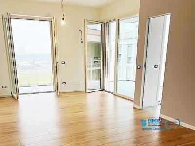 Stanza vuota con pavimento in legno, due porte aperte che conducono ad un balcone e una porta chiusa sul lato, ideale per le pulizie post-costruzione (pulizie di fine cantiere) a Milano