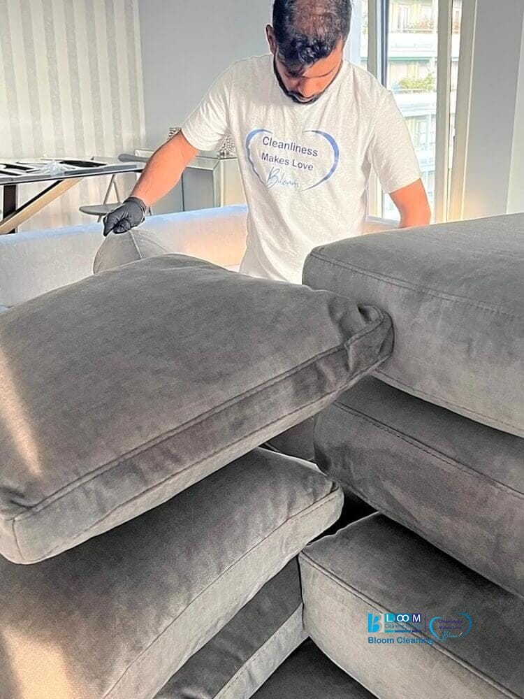 Una persona che indossa una maglietta bianca e guanti sta pulendo meticolosamente una pila di grandi cuscini grigi in una stanza interna, esemplificando il servizio esperto fornito da Bloom Cleaning per il lavaggio divani domicilio Segrate.