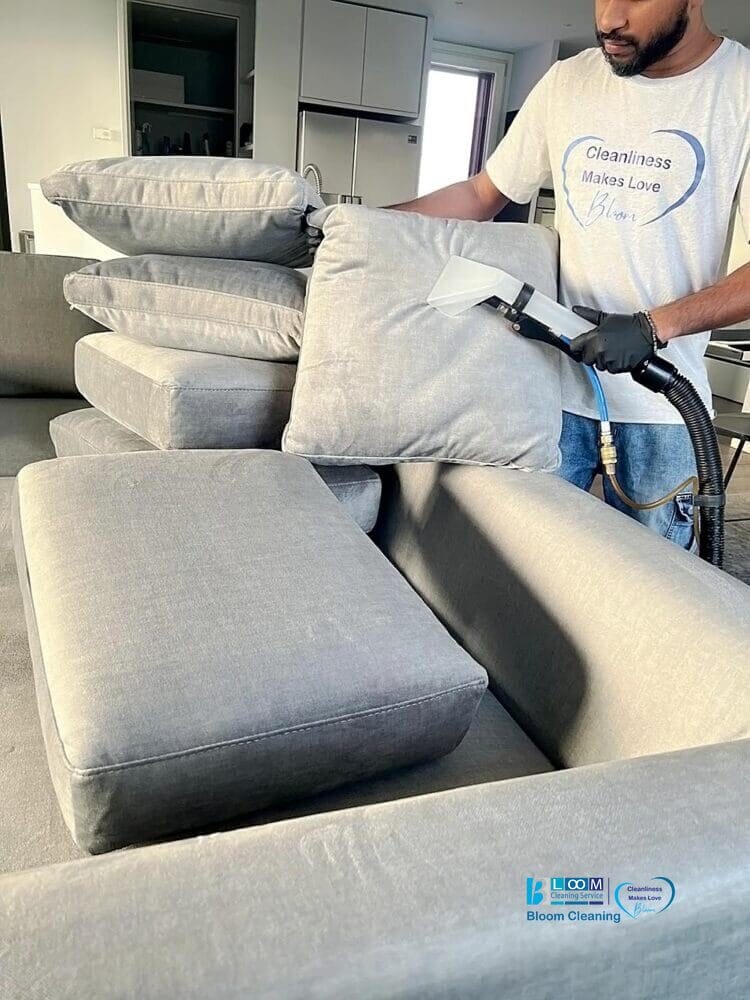 Una persona che indossa i guanti usa una macchina per la pulizia a vapore per pulire i cuscini di un divano grigio in un salotto moderno, a dimostrazione dell'eccezionale servizio di lavaggio divani domicilio Segrate di Bloom Cleaning.