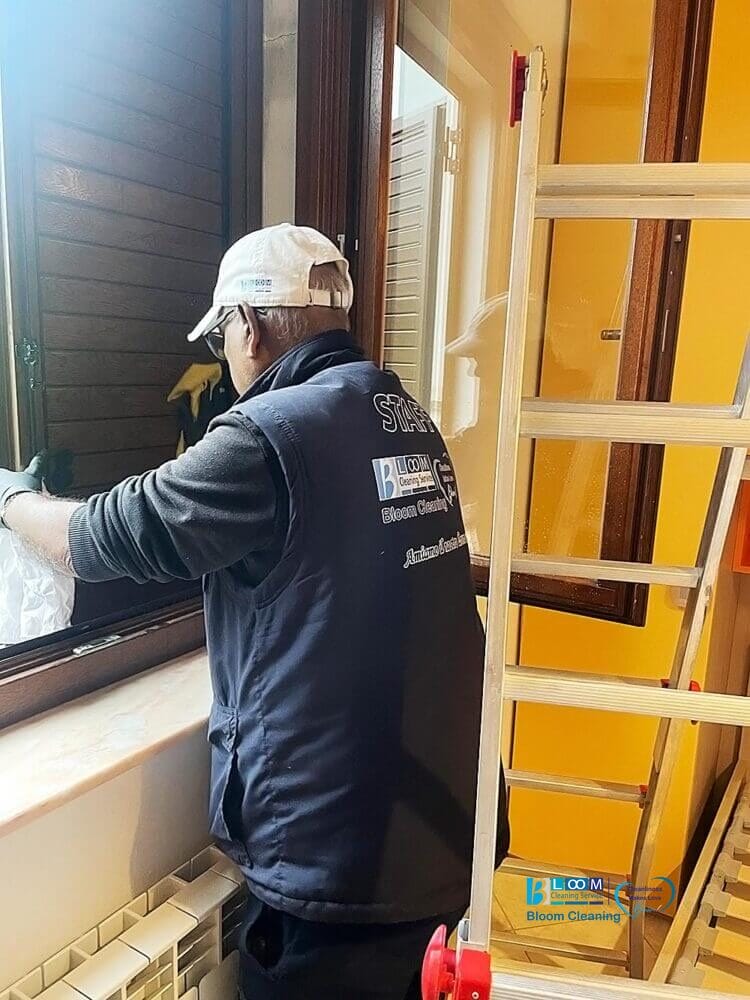 Una persona che indossa un berretto e un gilet di Bloom Cleaning impresa di pulizie sta pulendo una finestra accanto a una scala in una casa.