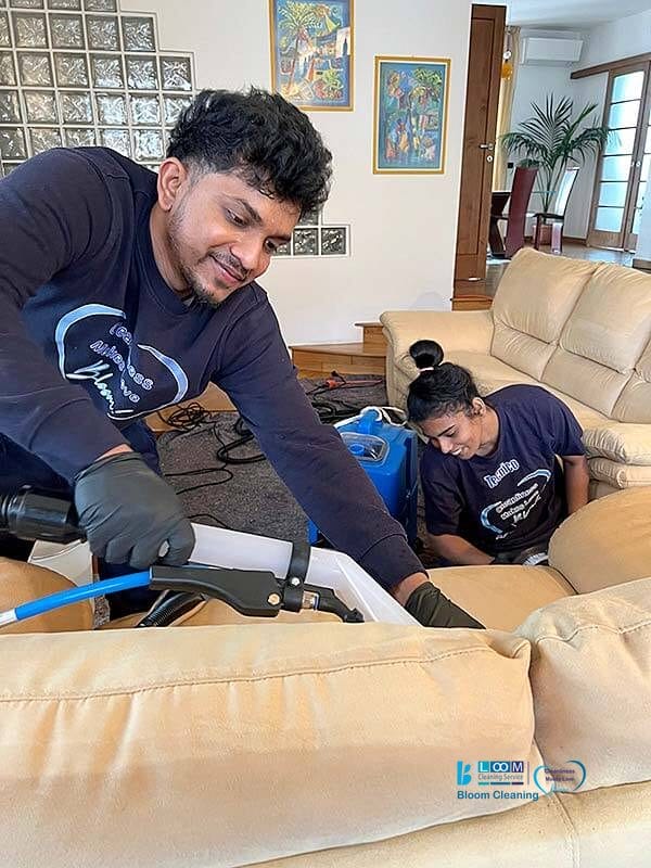 Due persone in uniforme di Bloom Cleaning stanno pulendo un divano beige in un soggiorno utilizzando attrezzature professionali. Sorridono e sembrano concentrati sul loro lavoro, mettendo in mostra l'eccellenza di Pulizia Divani a Domicilio a Paderno Dugnano.