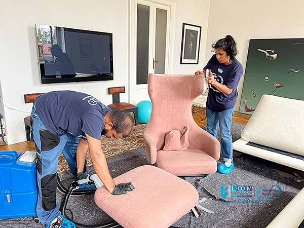 Due persone in uniforme di Bloom Cleaning stanno pulendo meticolosamente una poltrona rosa e il suo pouf in un soggiorno con TV montata e mobili moderni. Uno utilizza uno strumento di pulizia, mentre l'altro si concentra sui dettagli della sedia, garantendo una Pulizia Divani a Domicilio a Cinisello Balsamo di prim'ordine.