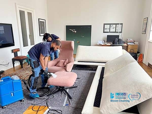 Due persone di Bloom Cleaning stanno pulendo una sedia imbottita rosa in un soggiorno, utilizzando una macchina per la pulizia. La stanza ha un divano bianco, una TV e vari mobili e decorazioni. Pulizia Divani a Domicilio a Cinisello Balsamo assicura che la tua casa sia al meglio!