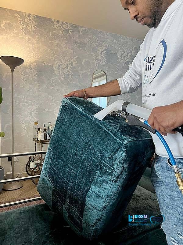 Un uomo con una felpa bianca pulisce un cuscino verde acqua utilizzando uno strumento di pulizia collegato a un tubo blu in una stanza arredata con stile, dimostrando gli efficienti servizi di Pulizia Divani a Domicilio a Lainate di Bloom Cleaning.