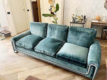 Un divano in velluto verde acqua con tre cuscini di seduta e cuscini per lo schienale è collocato in un elegante soggiorno a Lainate, completato da un carrello bar e una pianta sullo sfondo. Il pavimento in legno aggiunge calore, mentre le pareti dai colori chiari illuminano lo spazio. I servizi di Pulizia Divani garantiscono che rimanga immacolata.