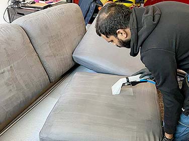 In una stanza di Peschiera Borromeo, una persona con una felpa nera utilizza una macchina portatile per la pulizia dei divani, pulendo meticolosamente un cuscino grigio del divano.