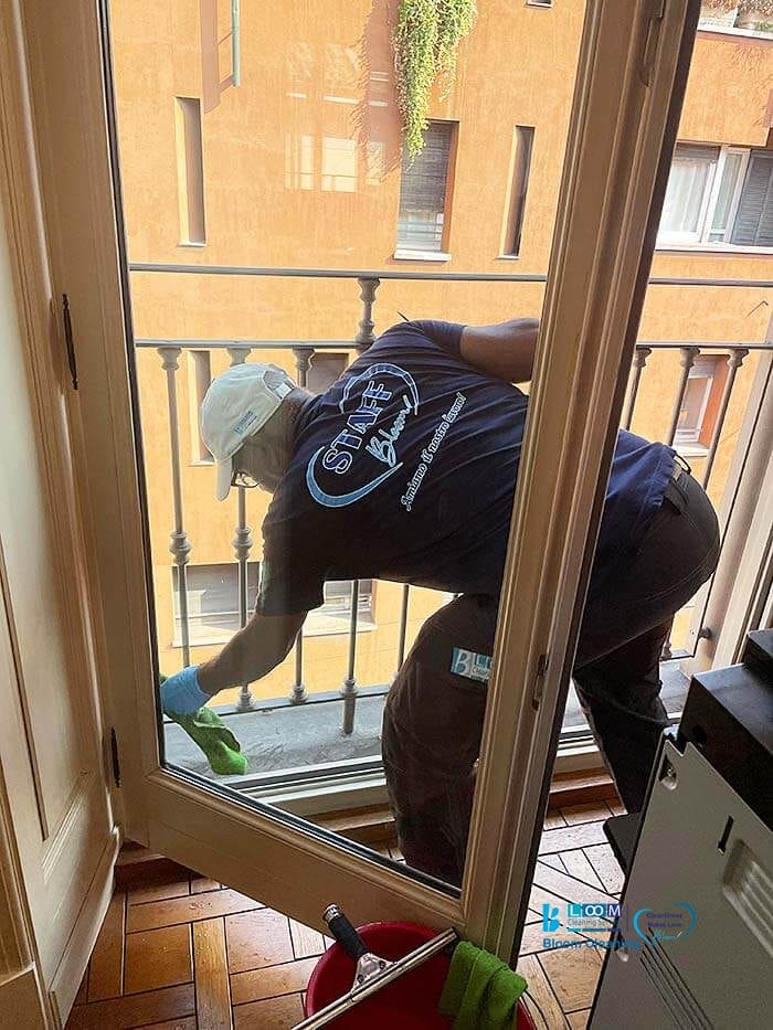Una persona che indossa un berretto e dei guanti sta pulendo l'esterno di una finestra sporgendosi parzialmente da essa, con vista su un balcone e un edificio color marrone chiaro. Bloomcleaning offre risultati impeccabili anche ad altezze difficili.