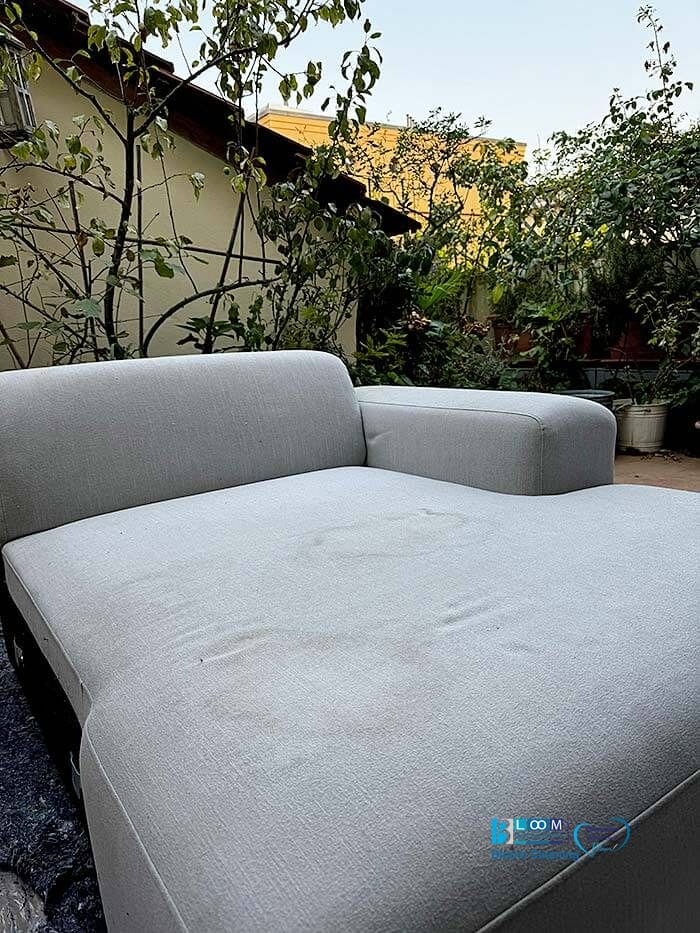 Una chaise longue da esterno grigio chiaro con macchie visibili è posizionata su un patio circondato dal verde e da un edificio sullo sfondo. Considera Pulizia divani domicilio a Buccinasco per ripristinare le sue condizioni immacolate in questo ambiente sereno.