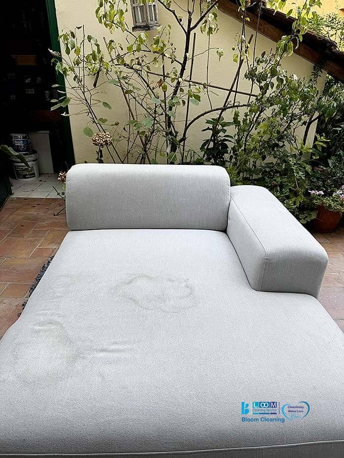 Una chaise longue da esterno grigio chiaro con visibili macchie d'acqua è posizionata su un patio piastrellato vicino a piante e verde, perfetta per Pulizia divani domicilio a Buccinasco.