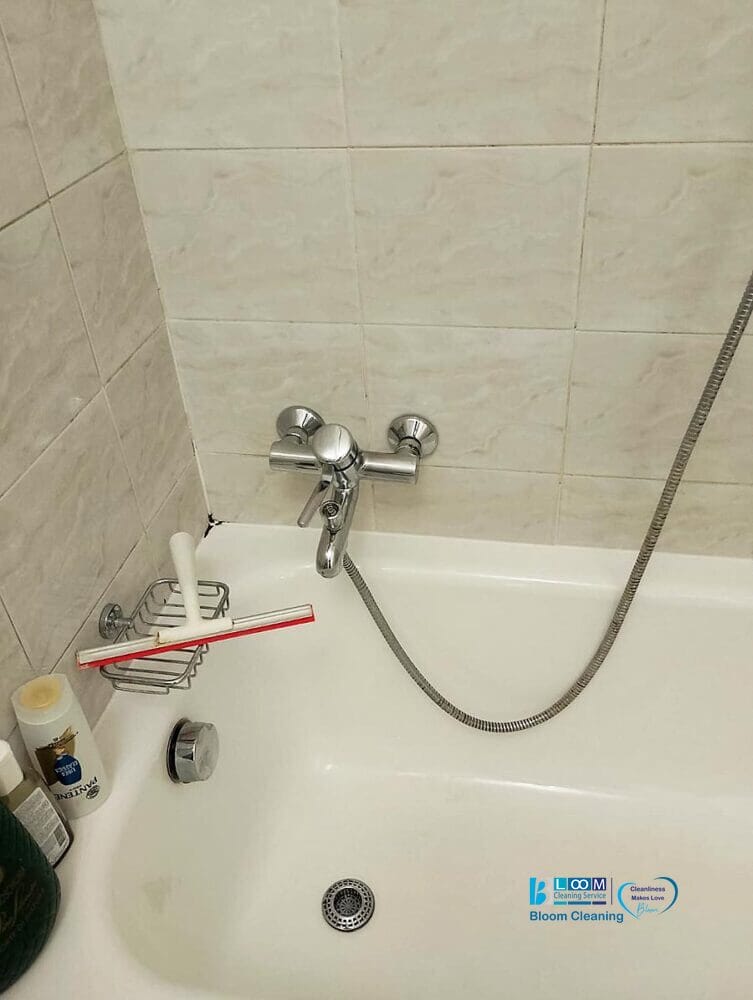 Una vasca da bagno con un rubinetto a parete e una doccetta portatile. C'è un tergivetro rosso appeso a un portasapone e vari articoli da toeletta sono posizionati sul bordo della vasca, assicurando pulizia bagno per un risultato sorprendente.