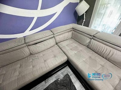 Il divano componibile grigio con cuscini trapuntati è appoggiato elegantemente contro una parete decorata con motivi geometrici magenta e bianchi. Una lampada da terra illumina dolcemente l'angolo, illuminando lo spazio invitante, perfetto per una giornata di Bloom Cleaning o per la routine di Pulizia Divani.