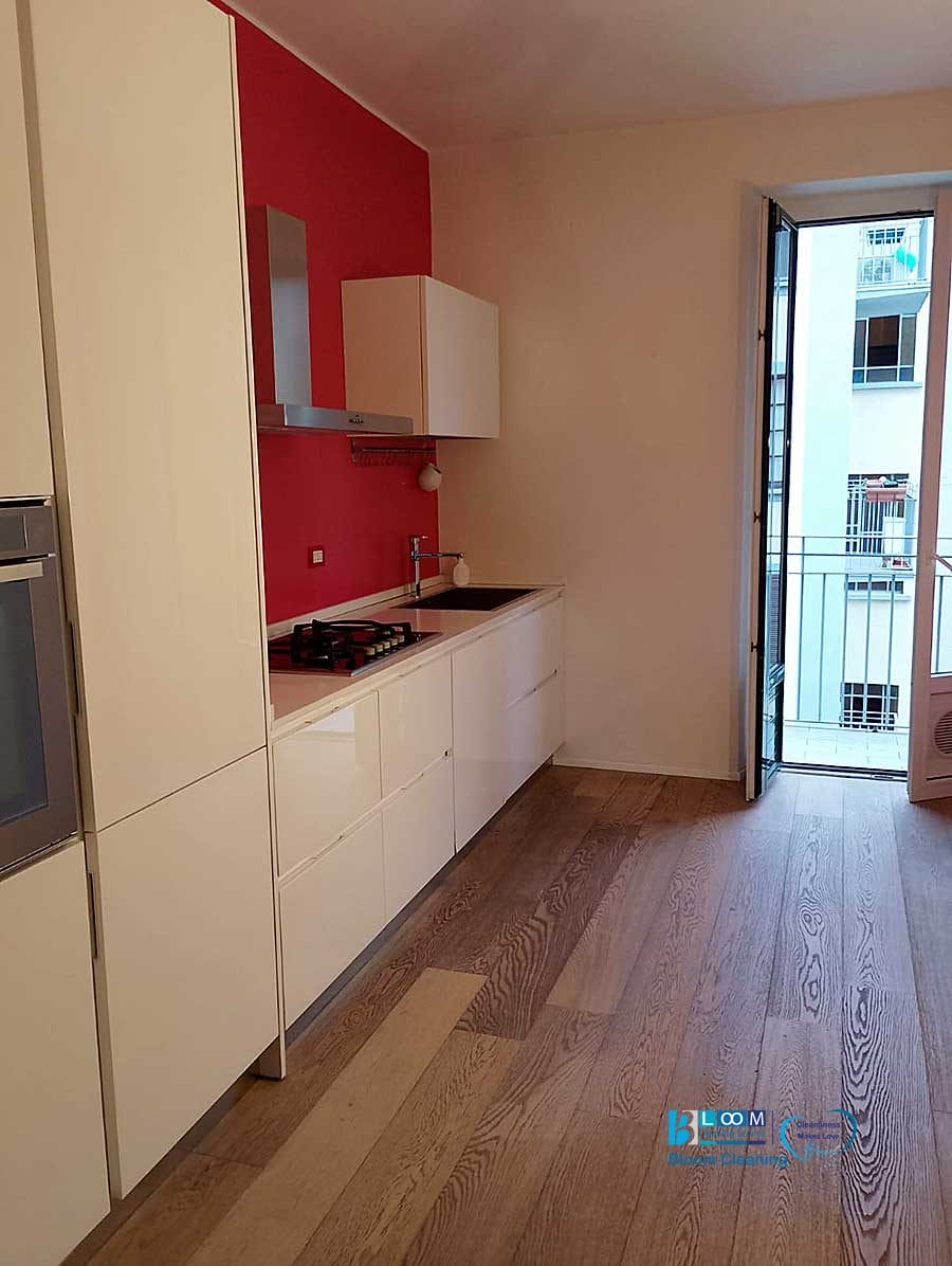 Cucina moderna con mobili bianchi, un'elegante stufa a gas e una parete decorativa rossa. Il design Bozza si fonde in modo fluido con il pavimento in legno che conduce a un balcone che offre viste sugli edifici adiacenti.