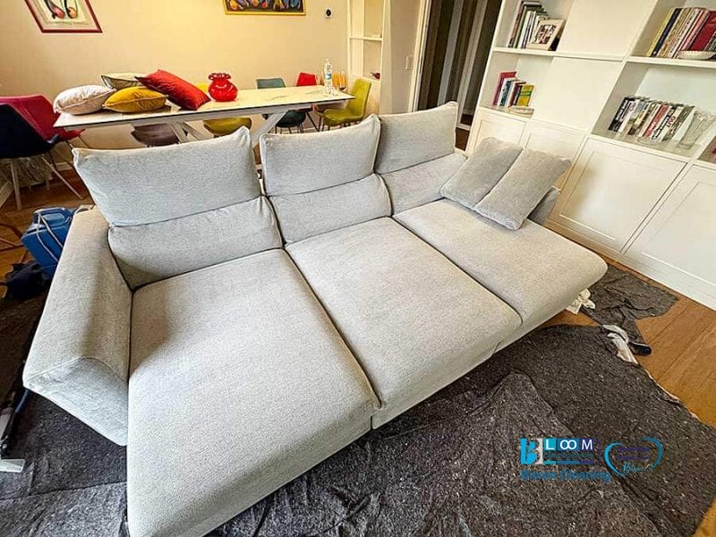 Un divano componibile grigio chiaro con sedili reclinabili è appoggiato su una fodera protettiva in un soggiorno, a dimostrazione del tocco impeccabile di Bloom Cleaning. La stanza è elegantemente arredata con un tavolo da pranzo e una libreria.
