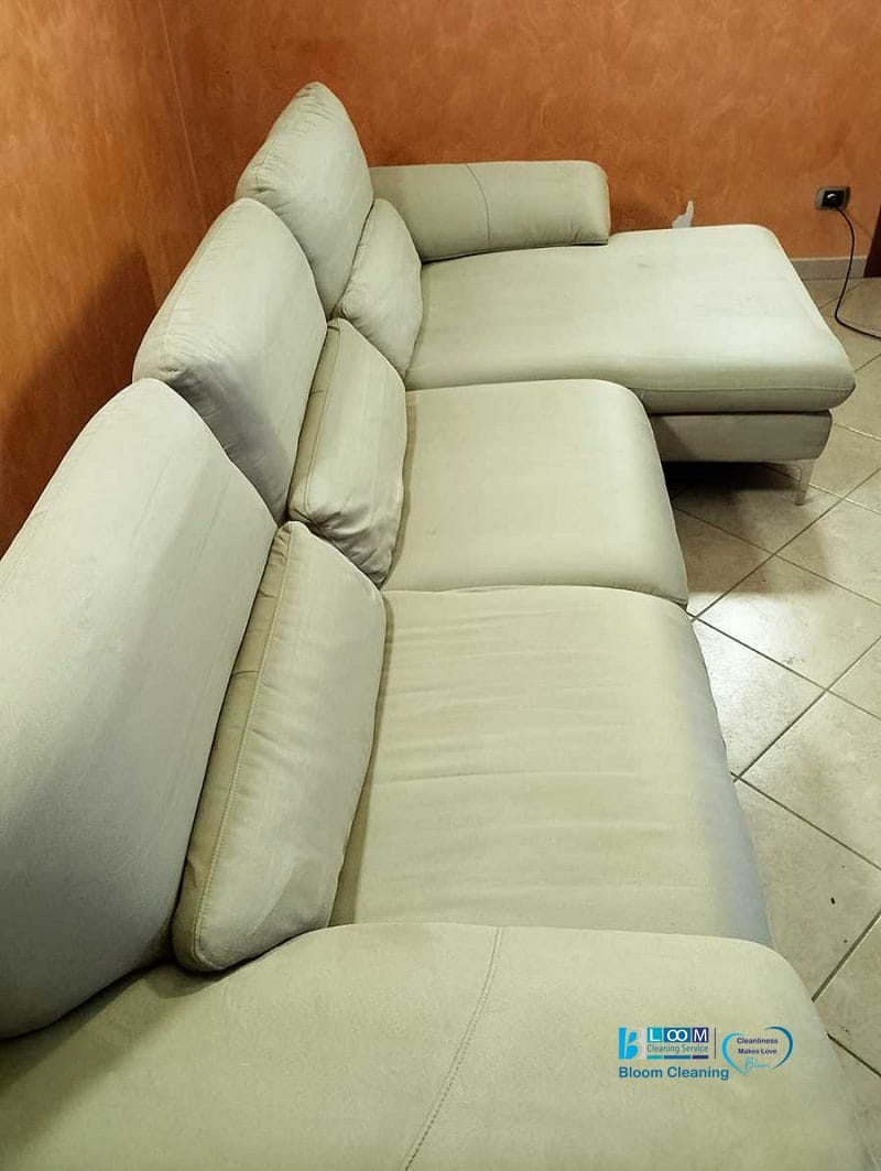 Un divano componibile beige con cuscini morbidi è sistemato in un angolo su un pavimento piastrellato, accanto a una parete arancione, creando uno spazio caldo e invitante. La disposizione sembra così naturale come se fosse stata progettata da bozza automatica.