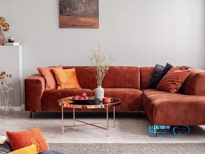 Un soggiorno moderno con un divano componibile color ruggine, cuscini decorativi e un tavolino rotondo in metallo presenta un dipinto murale. L'arredamento include piante secche in un vaso, tenute in perfette condizioni da Bloom Cleaning Service.