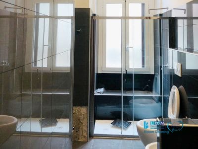 Un bagno moderno in un elegante Appartamento con porte doccia in vetro, sanitari bianchi e grandi finestre smerigliate, che ricordano il design elegante del Giambellino Milano.
