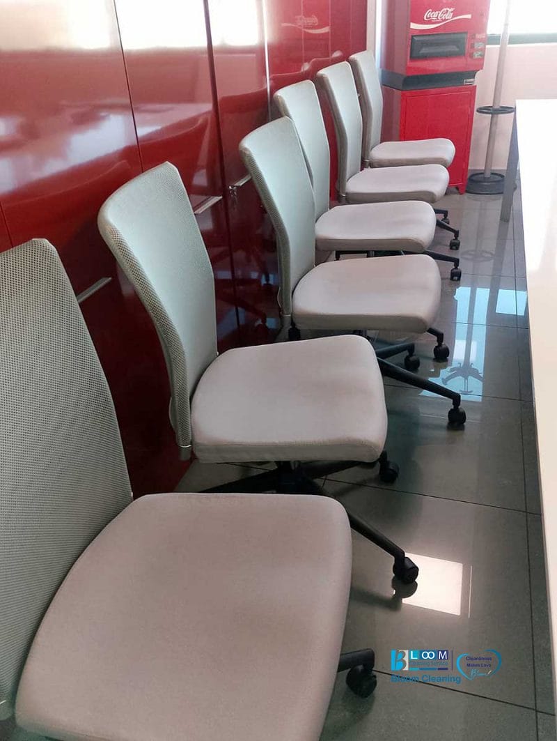 Cinque sedie da ufficio disposte ordinatamente in fila accanto a una parete rosso brillante riflettono l'eleganza di Ufficio Milano, con un distributore automatico di Coca-Cola visibile sullo sfondo. Le sedie mostrano i risultati di Pulizia Professionale Sedie per la massima pulizia e stile.