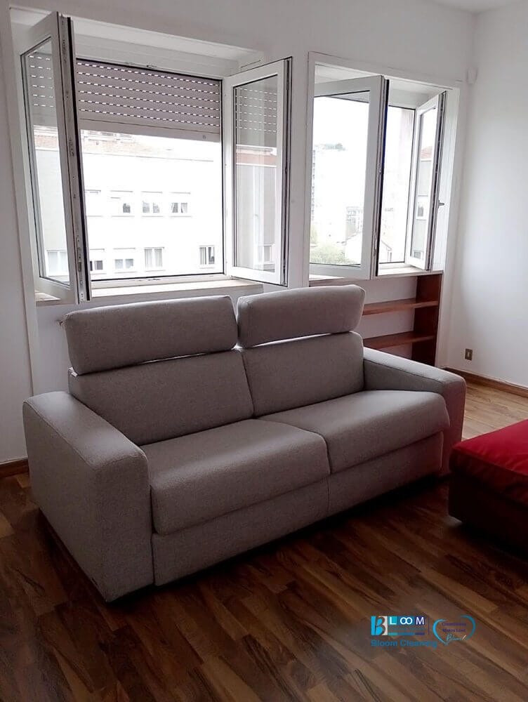Divano grigio in una stanza con pavimento in legno, posizionato di fronte a grandi finestre aperte. La luce naturale filtra, creando un ambiente sereno, mentre un piccolo mobile rosso aggiunge un tocco di eleganza. È come entrare nella tua bozza automatica di perfezione nell'interior design.