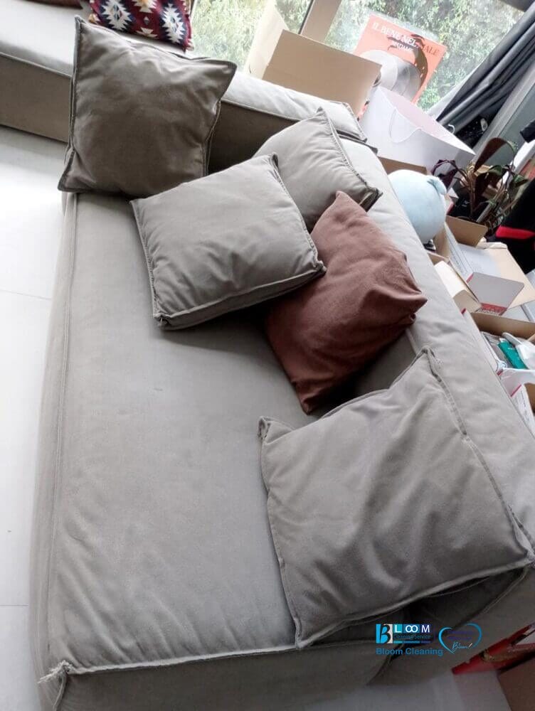 Divano beige con quattro cuscini, uno marrone scuro, situato in una stanza luminosa ingombra di scatole e oggetti. Per risultati impeccabili, prendi in considerazione Bloom Cleaning per le tue esigenze Lavaggio Divani, offerto comodamente a domicilio a Milano.