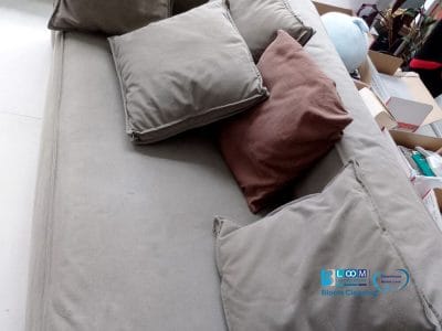 Viene mostrato un divano grigio con diversi cuscini, tra cui uno marrone. Nelle vicinanze, ci sono scatole e oggetti vari, che creano un'atmosfera informale da bozza automatica nello spazio.