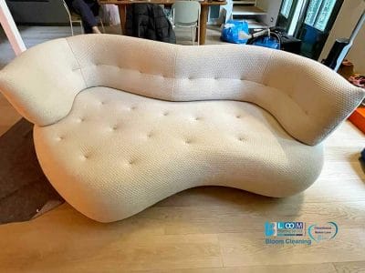 Un divano bianco curvo, di colore chiaro, con un motivo capitonné è appoggiato su un pavimento in legno in una stanza con un tavolo e delle sedie sullo sfondo.