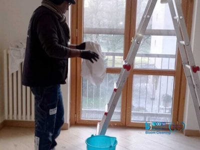 Una persona in piedi su un pavimento di legno tiene in mano uno straccio accanto a una scala e un secchio blu, preparandosi per una pulizia superficiale vicino alla grande finestra sullo sfondo.