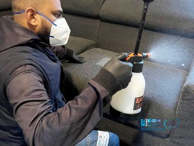 Indossando mascherina e guanti, una persona usa abilmente uno spruzzatore per rinfrescare il divano grigio con la precisione di una bozza automatica.
