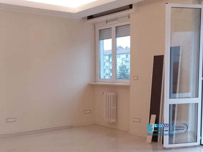 La stanza vuota, con le sue pareti bianche immacolate, una finestra con vista sugli edifici vicini e un radiatore sotto, sembra un appartamento vuoto. Una porta aperta si appoggia dolcemente contro il muro in questo spazio tranquillo situato in Zona Tolstoj Milano.