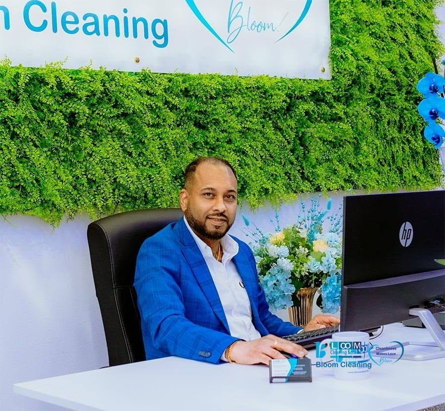 Manoj Perera è seduto a una scrivania con un computer e un mouse in mano. Dietro di lui, una parete verde con piante e un cartello con la scritta "Pulizia" e "Bloom Cleaning", che mette in risalto questa impresa di pulizie milanese.