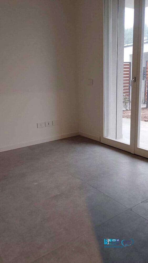 Stanza vuota in una Villetta ad Assago con ampio pavimento piastrellato, pareti bianche immacolate e un'alta porta a vetri che conduce all'esterno. Lo spazio risplende come nuovo dopo Pulizia Post-Cantiere, pronto per il suo prossimo capitolo di trasformazione.
