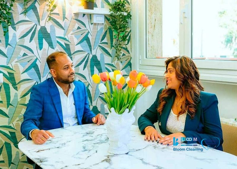 Due persone sedute a un tavolo di marmo con un vaso di tulipani, circondate da carta da parati a foglie e da una finestra. Entrambe indossano blazer blu e sono impegnate a conversare.