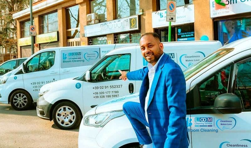Un uomo in abito blu sorride e si appoggia a un furgone bianco, con una fila di furgoni simili parcheggiati dietro di lui, accanto a un edificio.