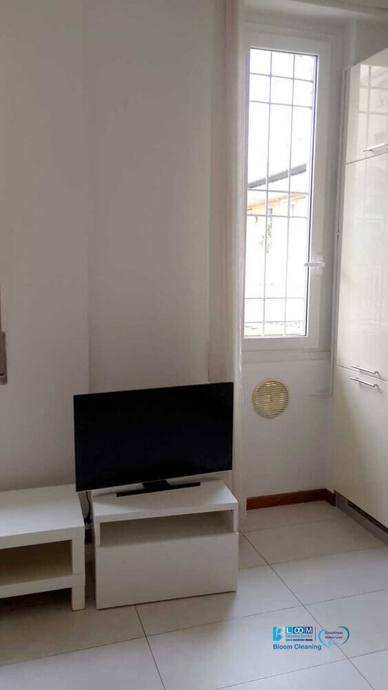 Una piccola TV a schermo piatto è appollaiata su un supporto bianco accanto a una finestra incorniciata da barre di metallo. Bozza automatica esalta la scena, evidenziando le pareti bianche e il pavimento piastrellato della stanza.