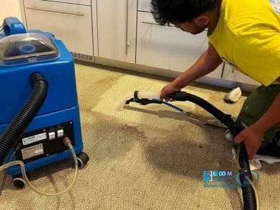 Una persona con una maglietta gialla sta utilizzando una macchina per la pulizia della moquette, occupandosi meticolosamente di una macchia sul tappeto nella zona della cucina.