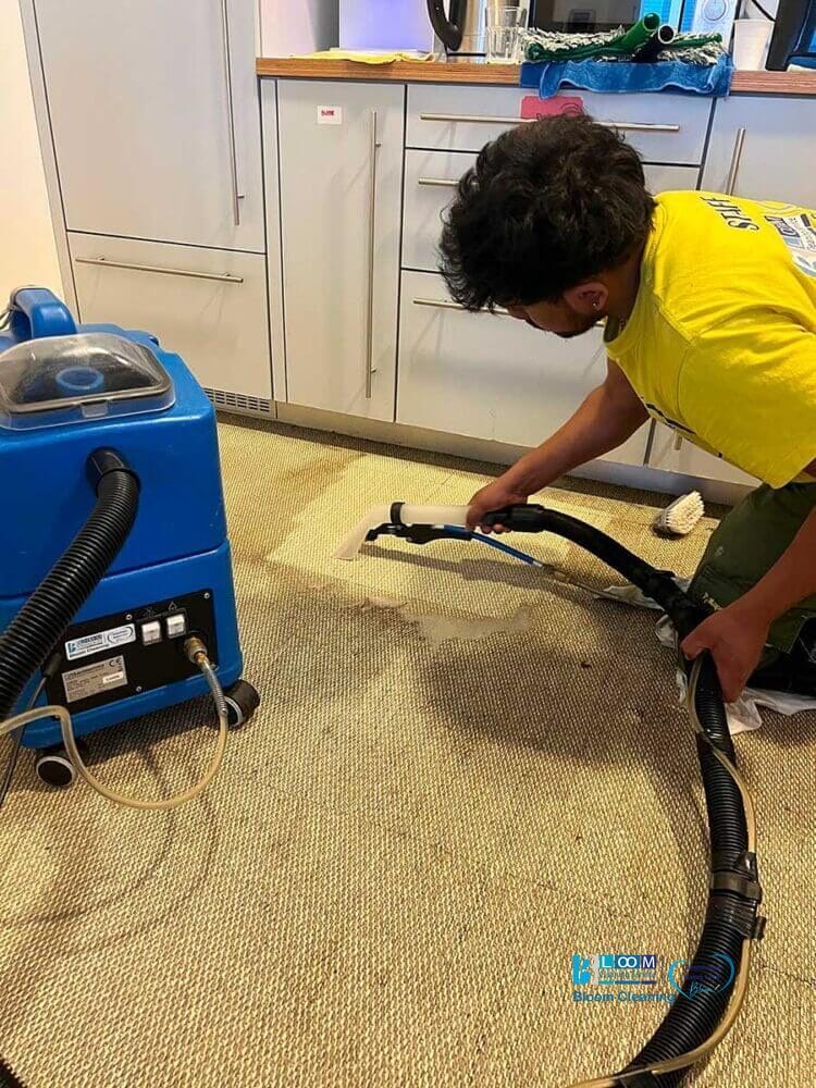 Una persona con una maglietta gialla sta utilizzando una macchina per la pulizia della moquette, occupandosi meticolosamente di una macchia sul tappeto nella zona della cucina.