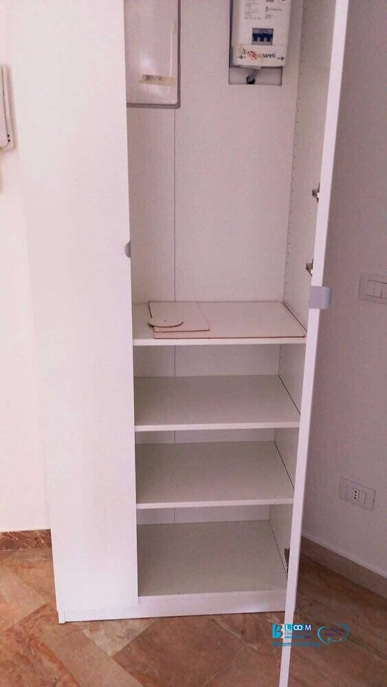 Un mobile bianco con quattro ripiani vuoti e un quadro elettrico sul ripiano superiore è appoggiato a una parete su un pavimento piastrellato: il contenitore ideale per i prodotti per la pulizia utilizzati dall'impresa di pulizie De Amicis Milano.