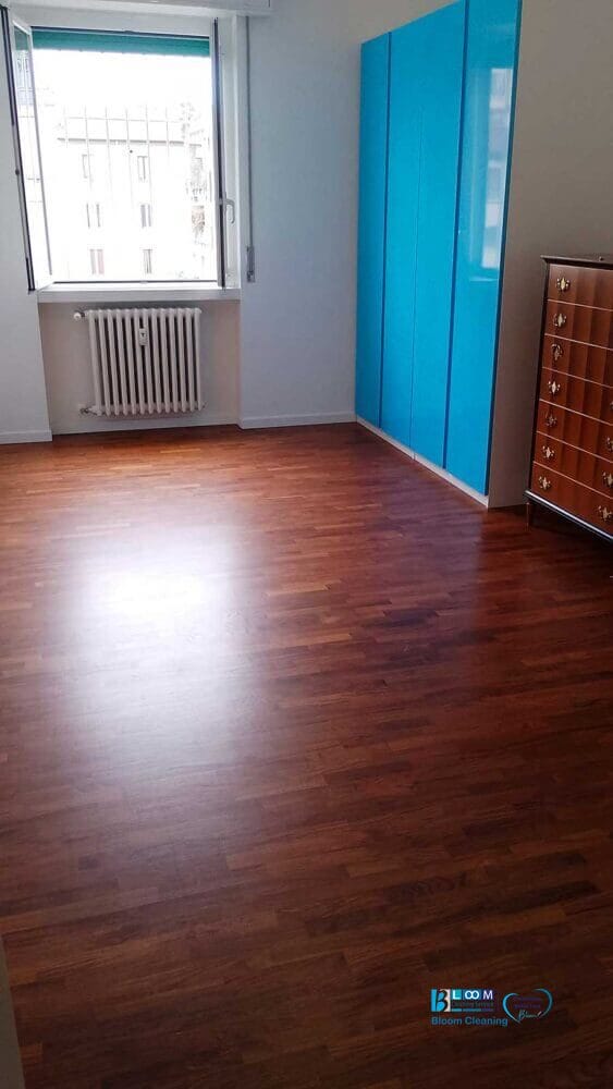 Stanza vuota con pavimento in legno, armadio blu, cassettiera in legno e un termosifone sotto una finestra con le sbarre: ideale per chi cerca pulizie appartamenti Milano o un'impresa di pulizie zona De Amicis Milano.