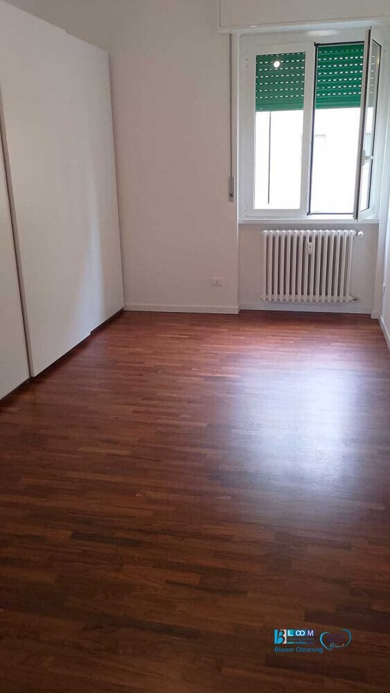 Stanza vuota con pavimento in legno, pareti bianche, un termosifone sotto una finestra con una persiana verde e un grande armadio bianco sulla sinistra: ideale per chi cerca pulizie appartamenti Milano per un nuovo inizio in uno spazio impeccabile.